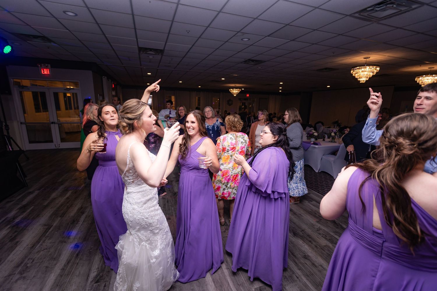 Bride dances joyfully with bridesmaids in purple gowns during lively wedding reception celebration.