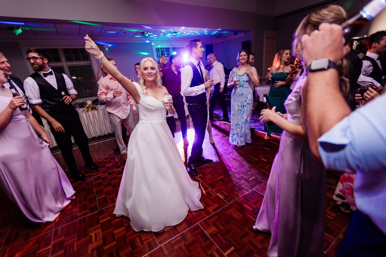 Bride in white gown raising her arm while dancing with guests under purple lights at a festive wedding reception at the Manor House.