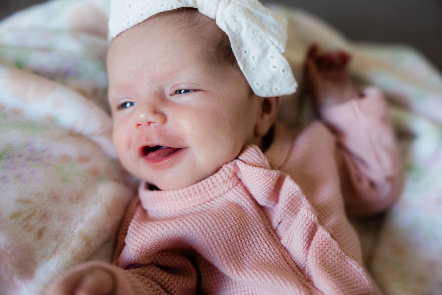 A smiling newborn baby girl in pink with a white bow headband lying on a soft blanket.