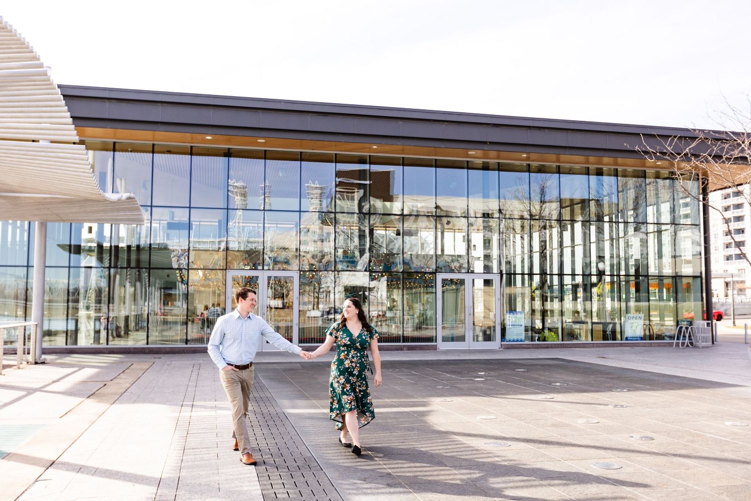 Couple holding hands and walking in front of a modern glass building on a sunny clear day.