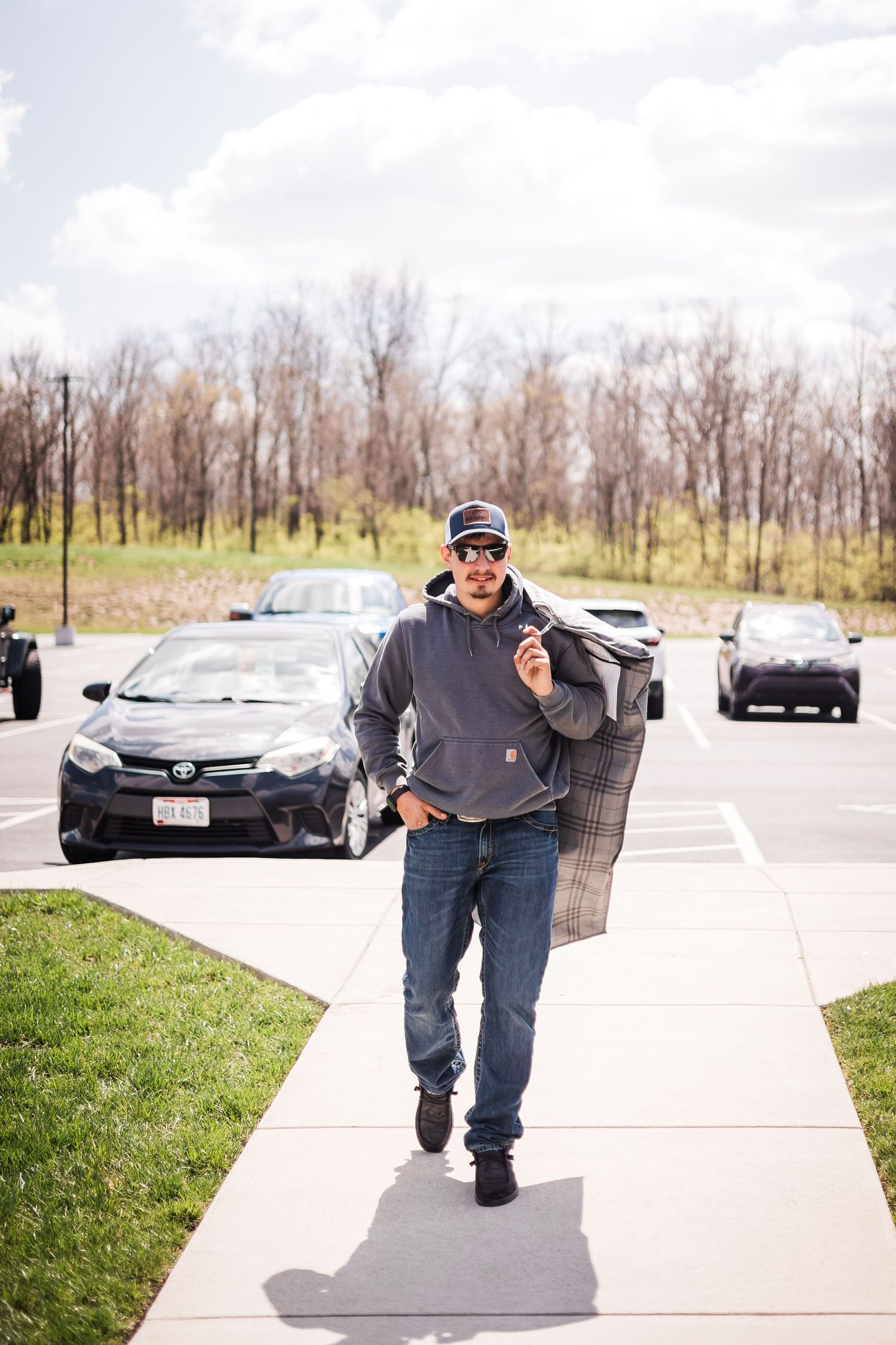 Grom in gray hoodie and jeans walks along sidewalk carrying jacket in parking lot.