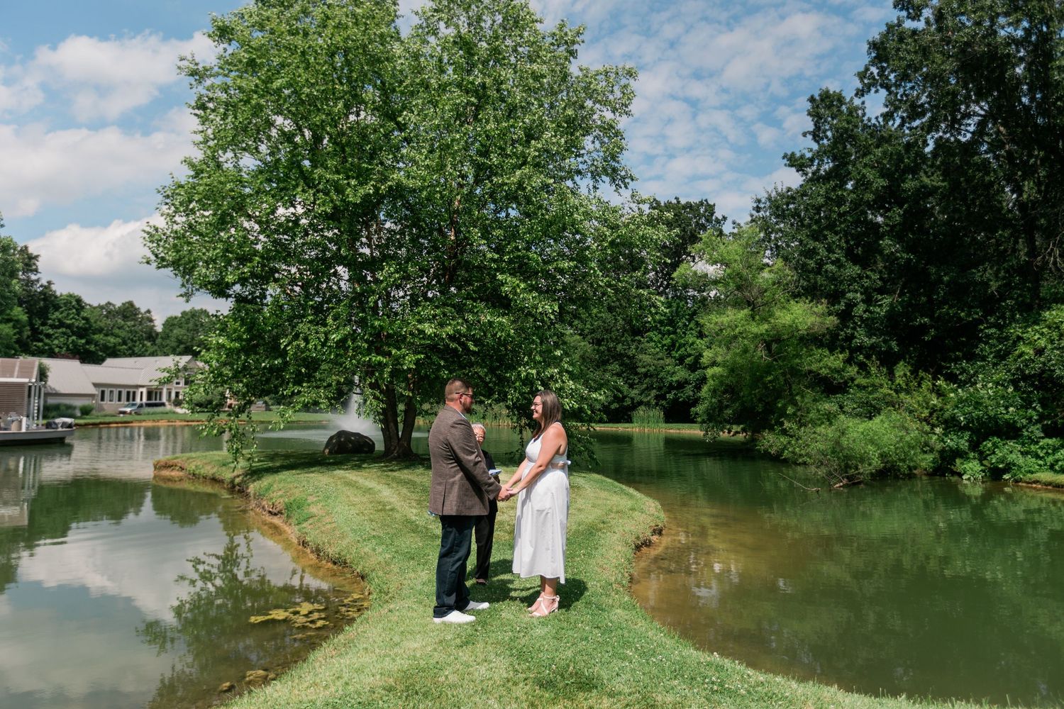 Couple sharing an intimate moment on a grassy peninsula surrounded by a peaceful pond during their wedding ceremony.