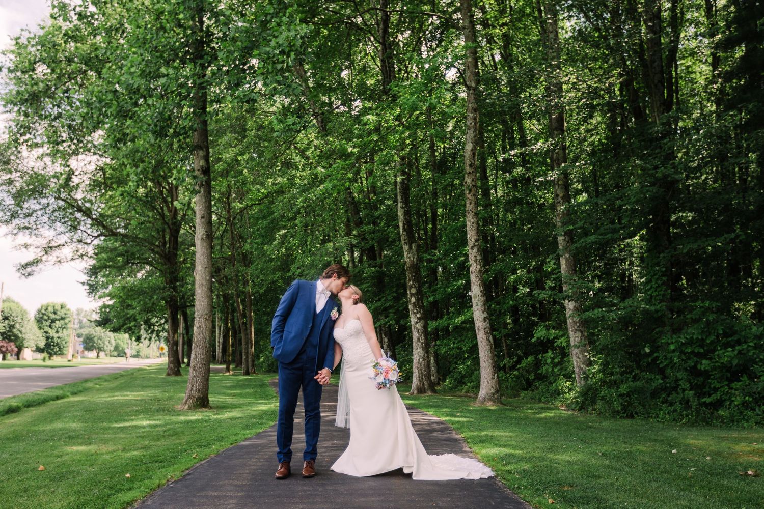 Bride and groom sharing a kiss on a tree-lined pathway surrounded by lush green trees.