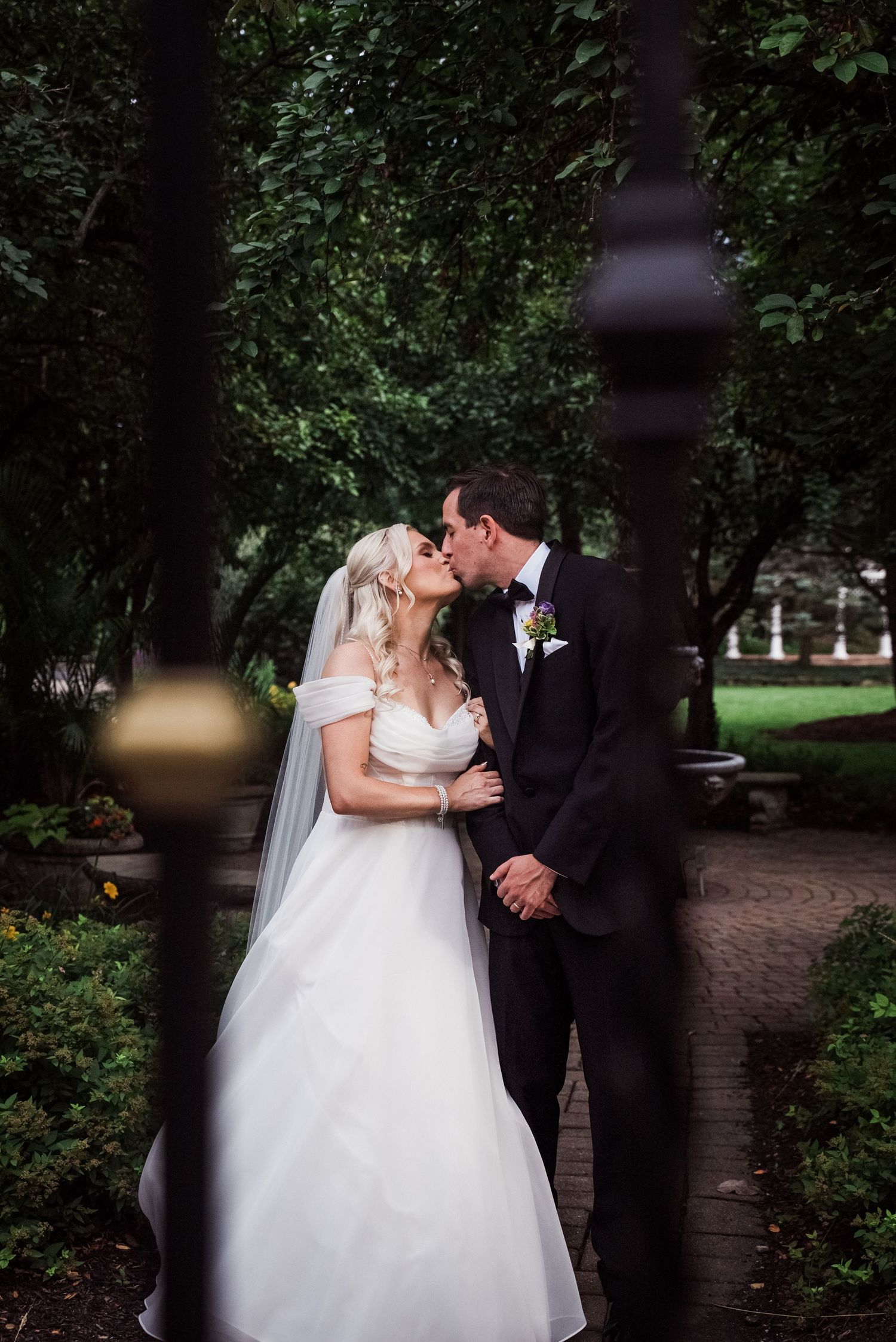 Bride and groom share a passionate kiss on a tree-lined garden pathway framed by dark iron fence posts.