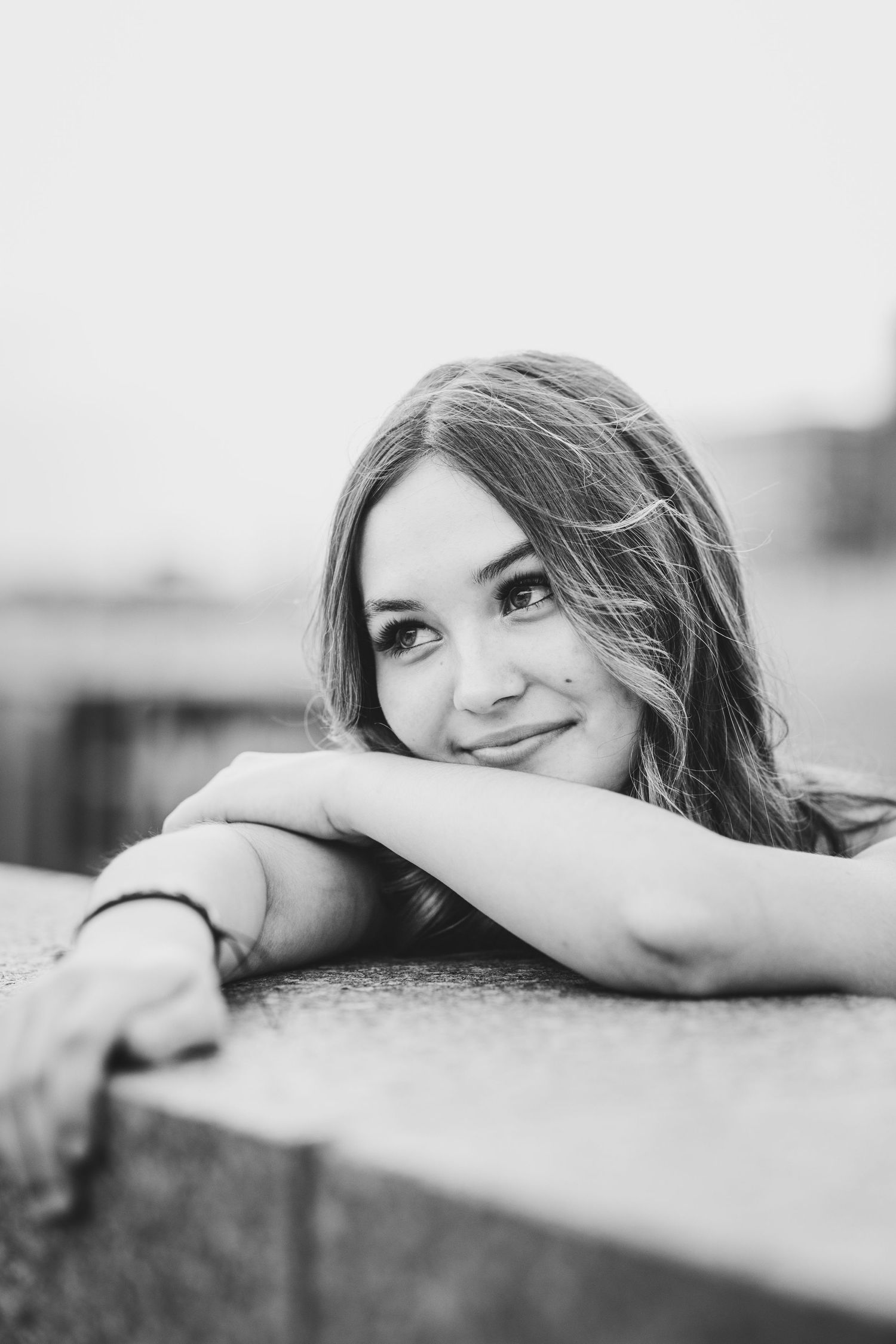 Close-up black and white portrait of a smiling young woman resting her chin on her arms outdoors.
