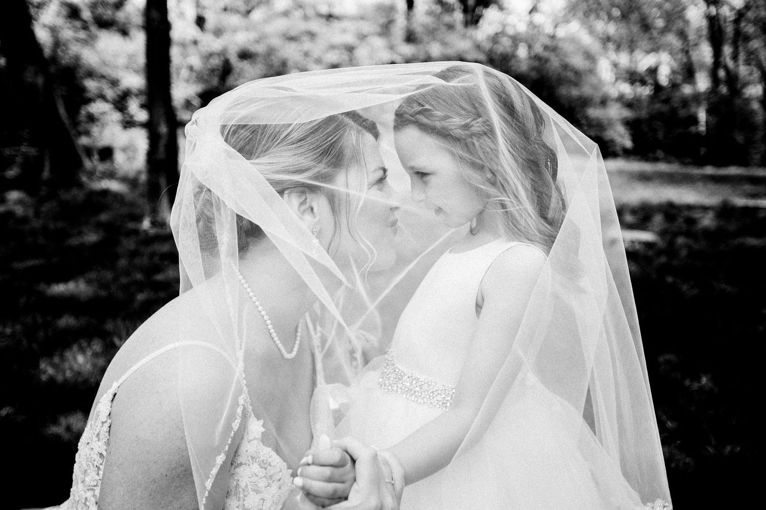 Black and white photo of two brides sharing a tender moment under a flowing veil outdoors.