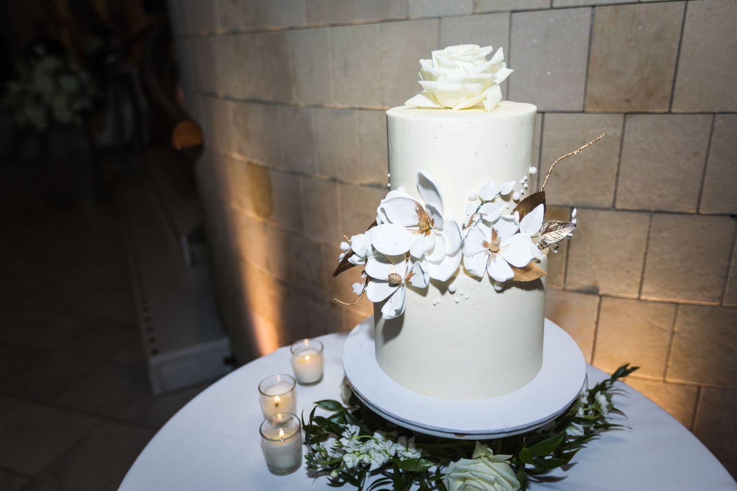 Elegant three-tier white wedding cake adorned with sugar flowers and a rose topper on a round table.