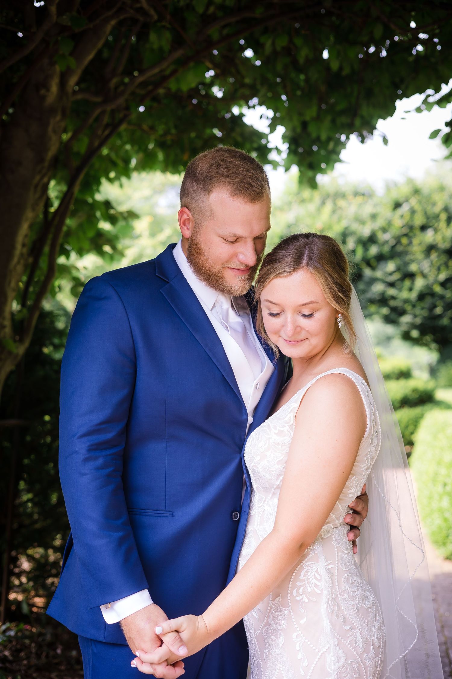 Bride and groom holding hands under lush green trees in a tender wedding portrait outdoors.
