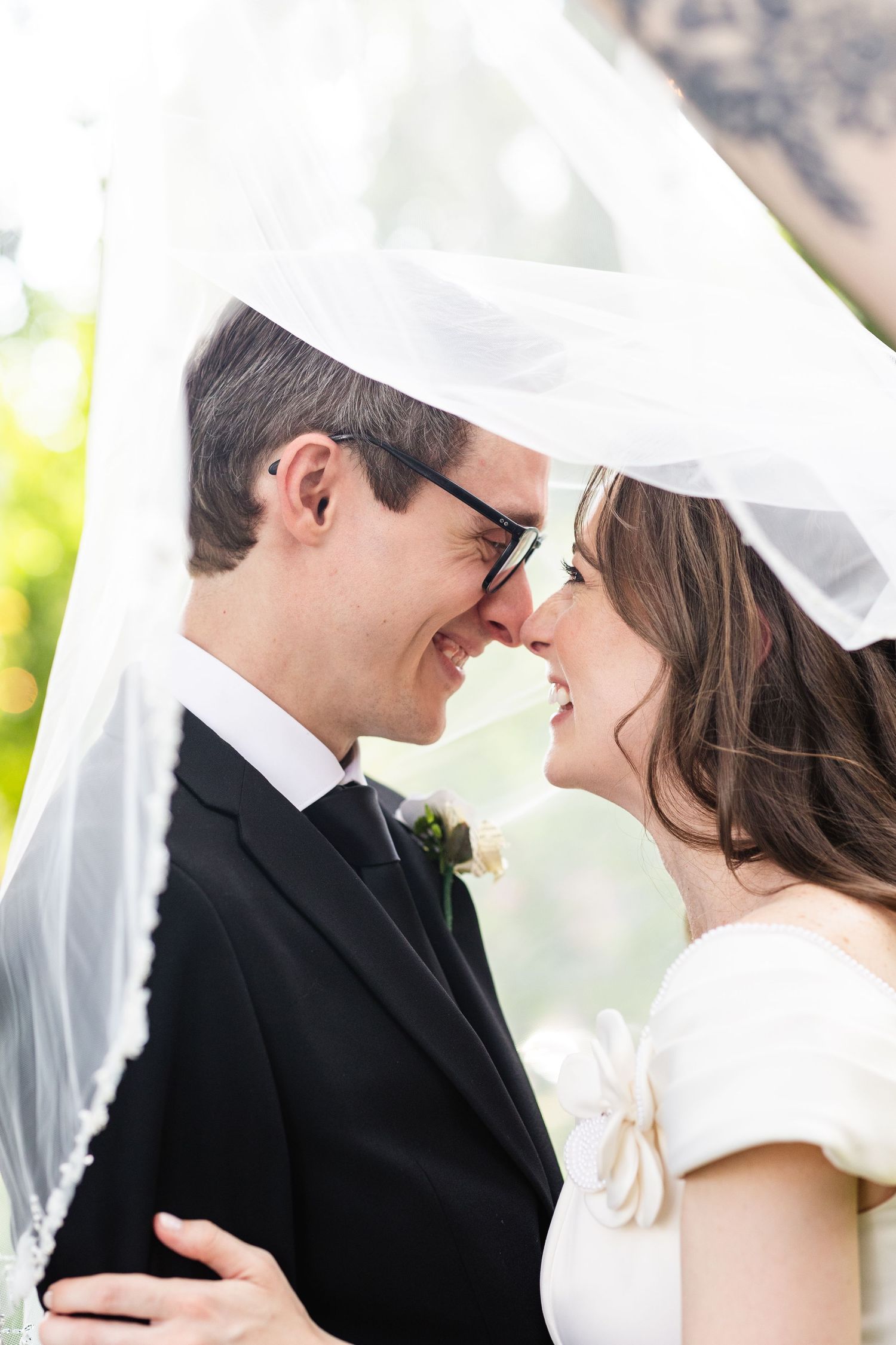 Laughing bride and groom touch noses under flowing white veil in intimate outdoor wedding portrait.
