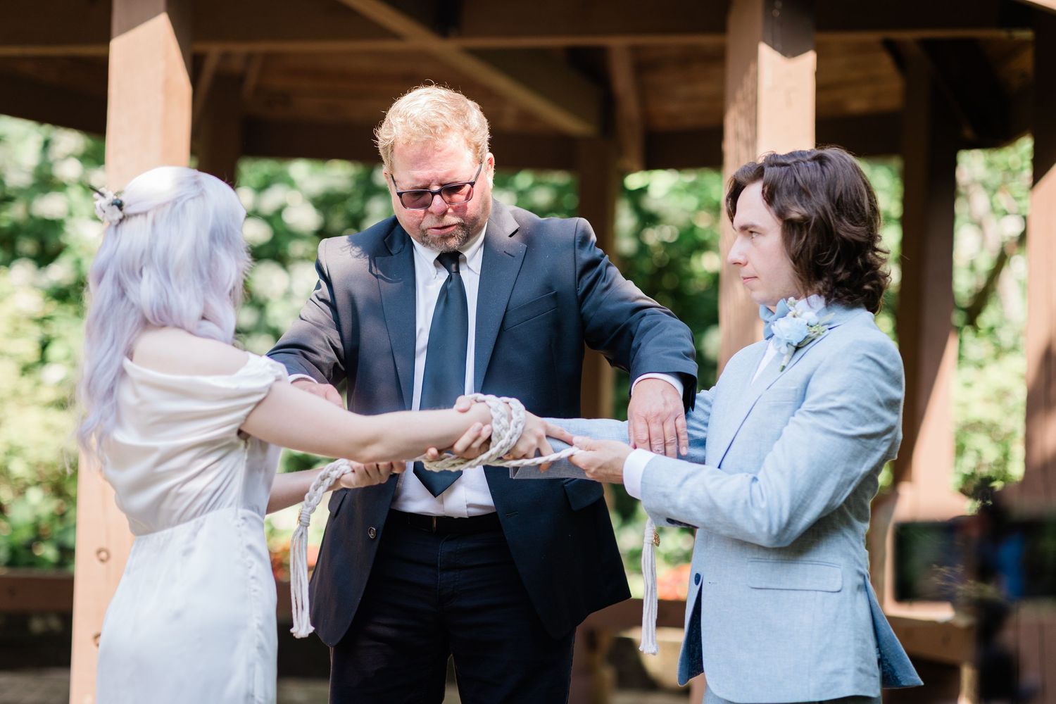 Wedding officiant in sunglasses ties handfasting rope around couple's hands during outdoor garden ceremony.