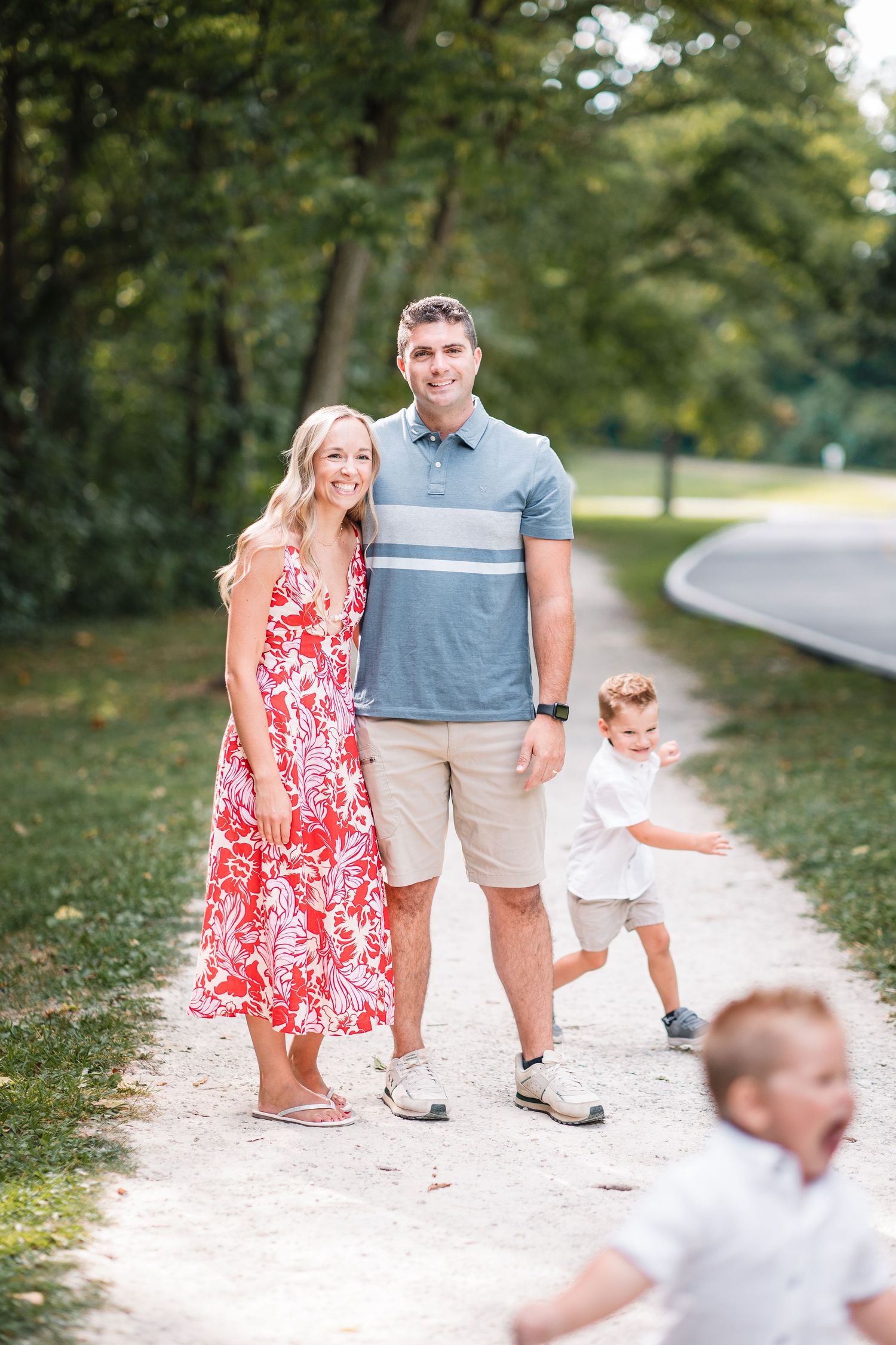 Smiling couple standing on a park path with two young boys playing nearby on a sunny day.