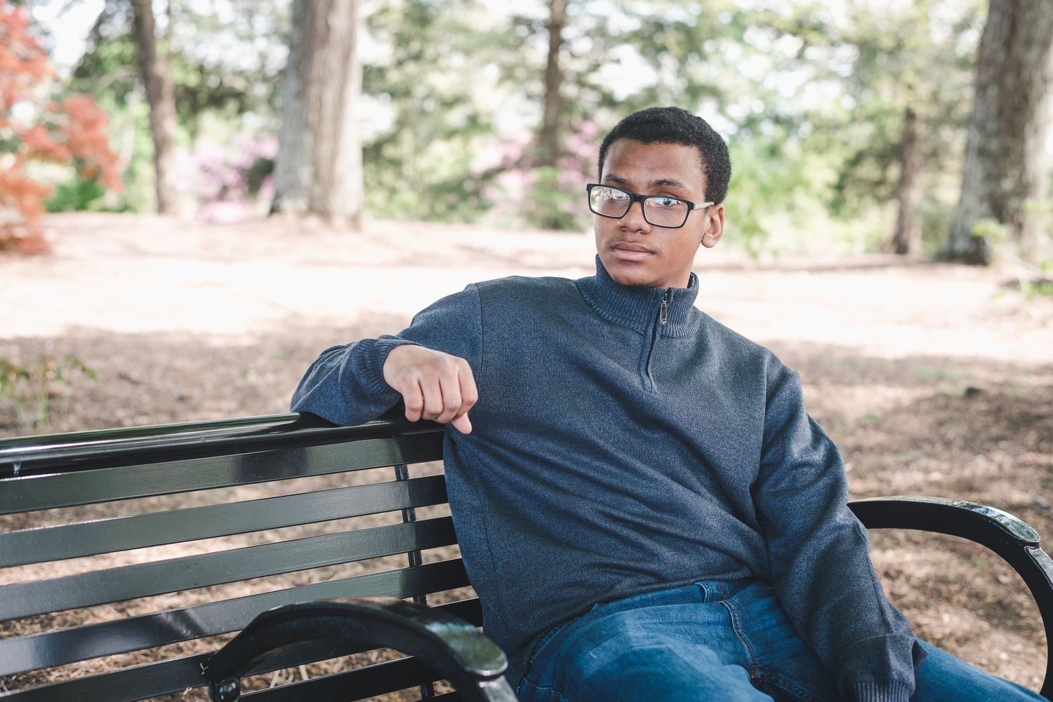 Young man in glasses and gray pullover sitting relaxed on park bench outdoors.
