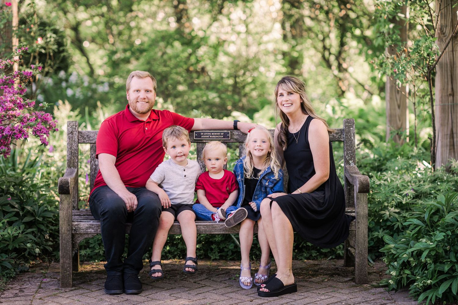 Family of five smiling on a wooden bench surrounded by colorful garden flowers.
