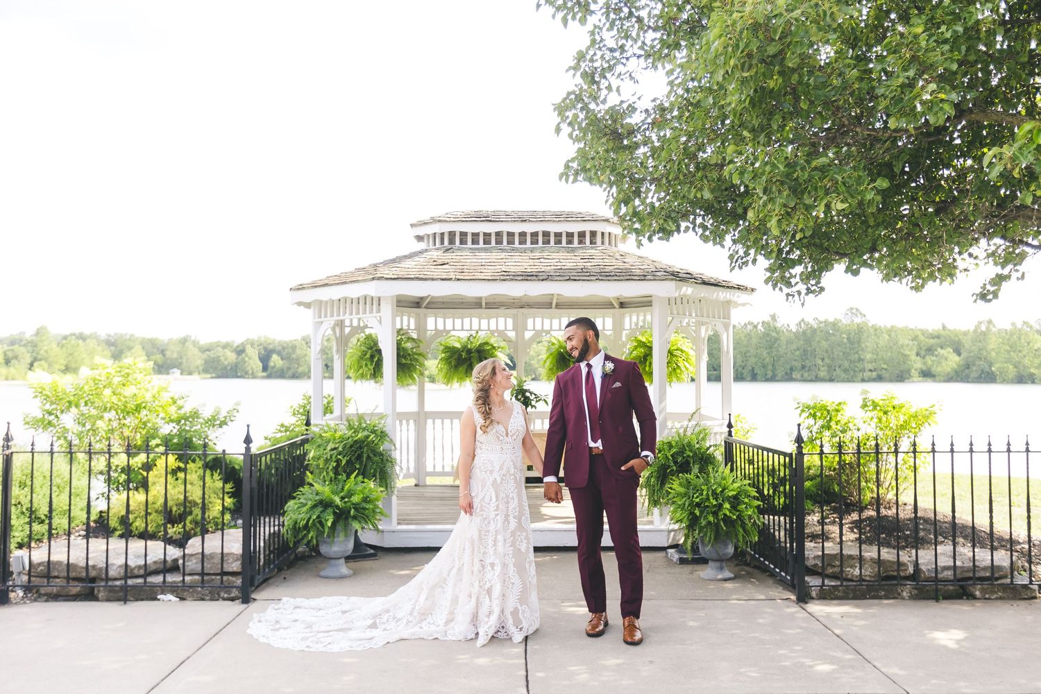 Newlywed couple holds hands near a white gazebo by a lake on their wedding day at Lake Lyndsay.