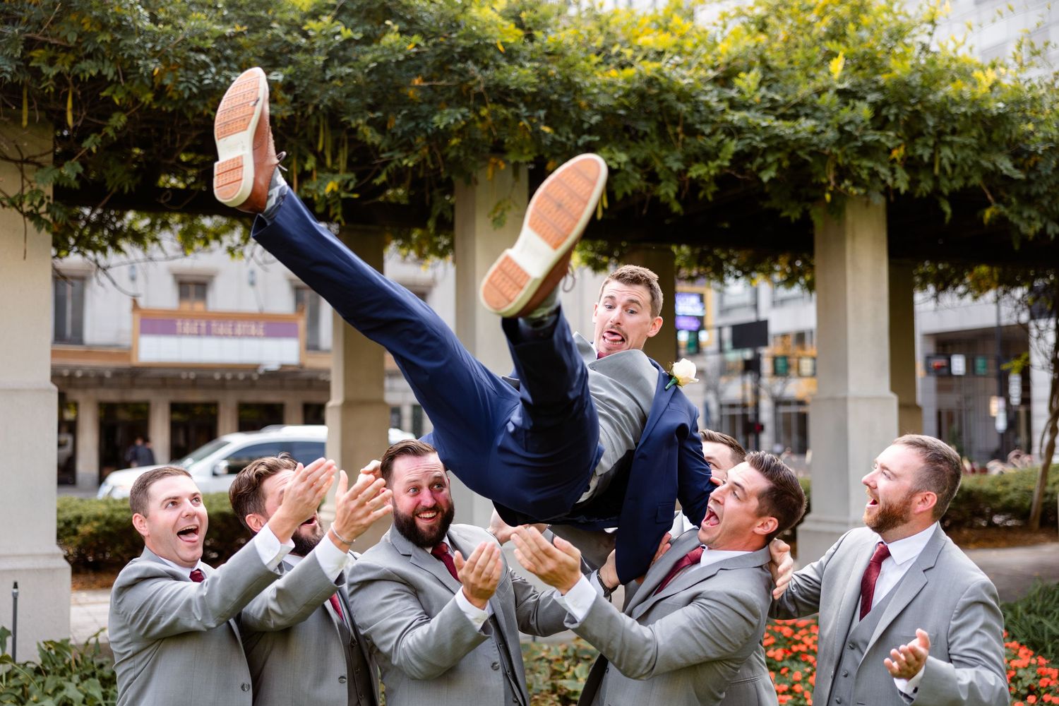Groomsmen in gray suits tossing groom in navy suit into the air outdoors during wedding celebration.