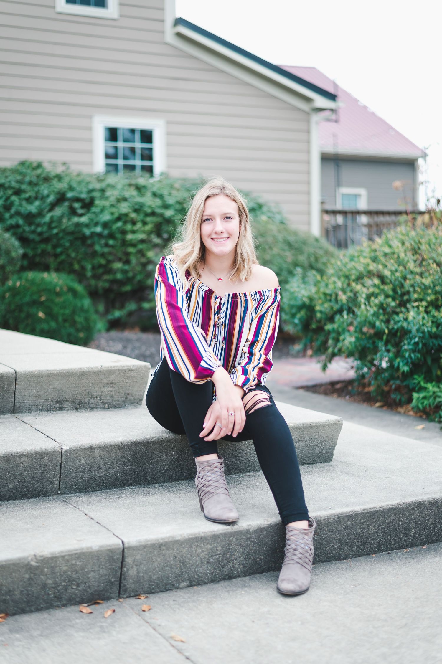 Smiling blonde teen sits on concrete steps wearing a striped off-shoulder top, black leggings, and boots.