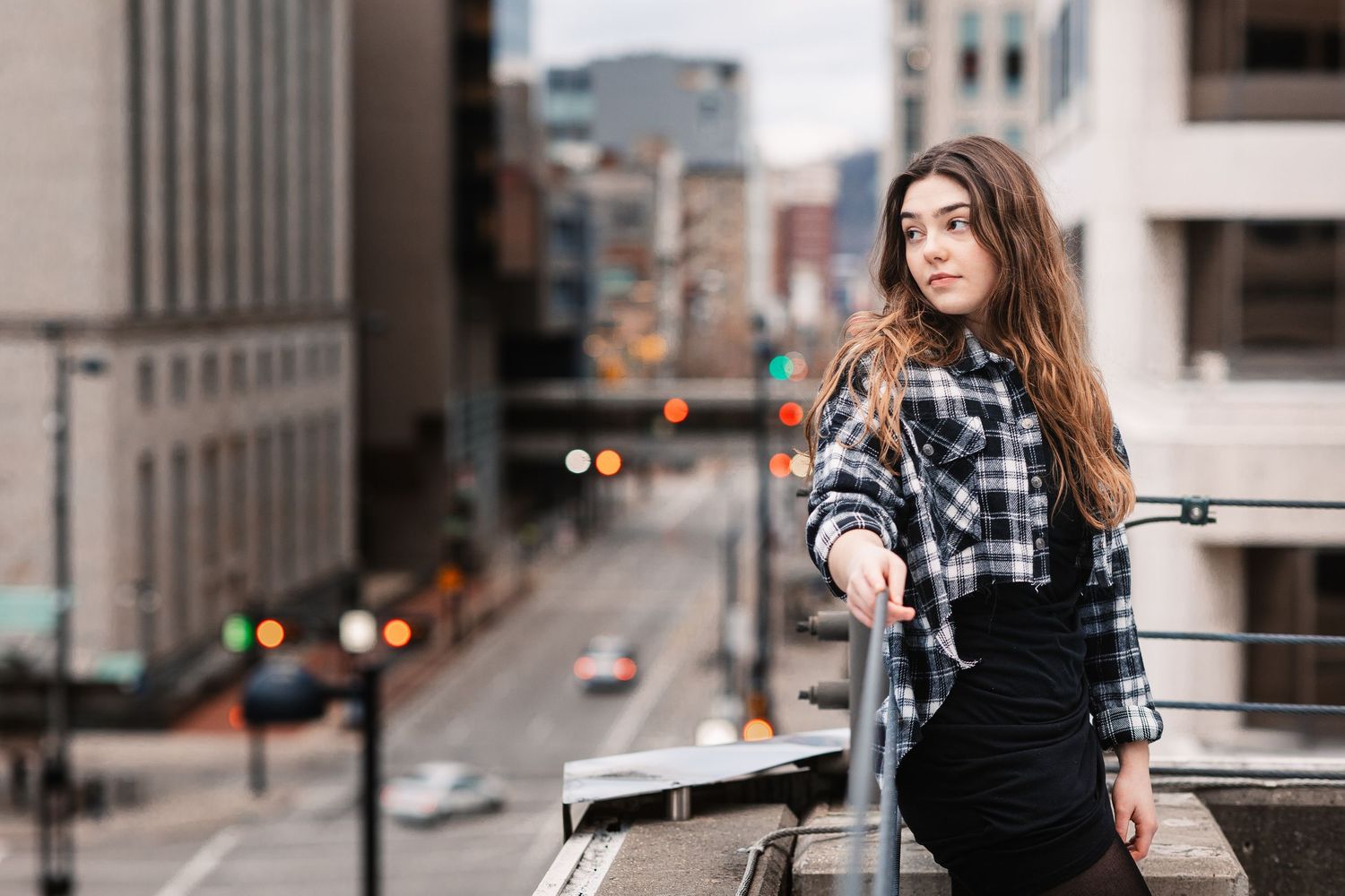 Young woman in plaid shirt leaning on railing with blurred city street background.