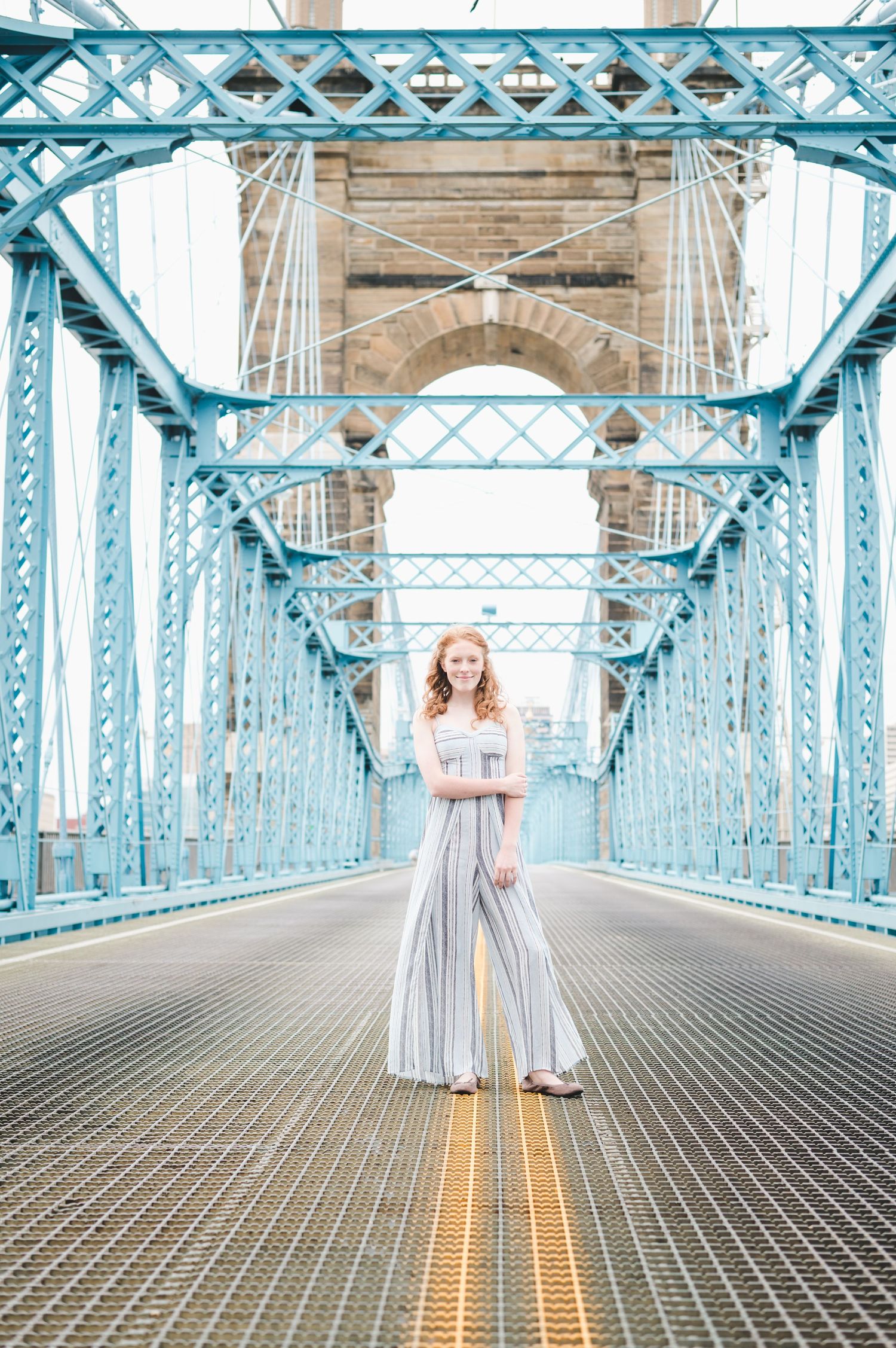 Young redhead woman poses on Roebling Bridge in a striped jumpsuit.