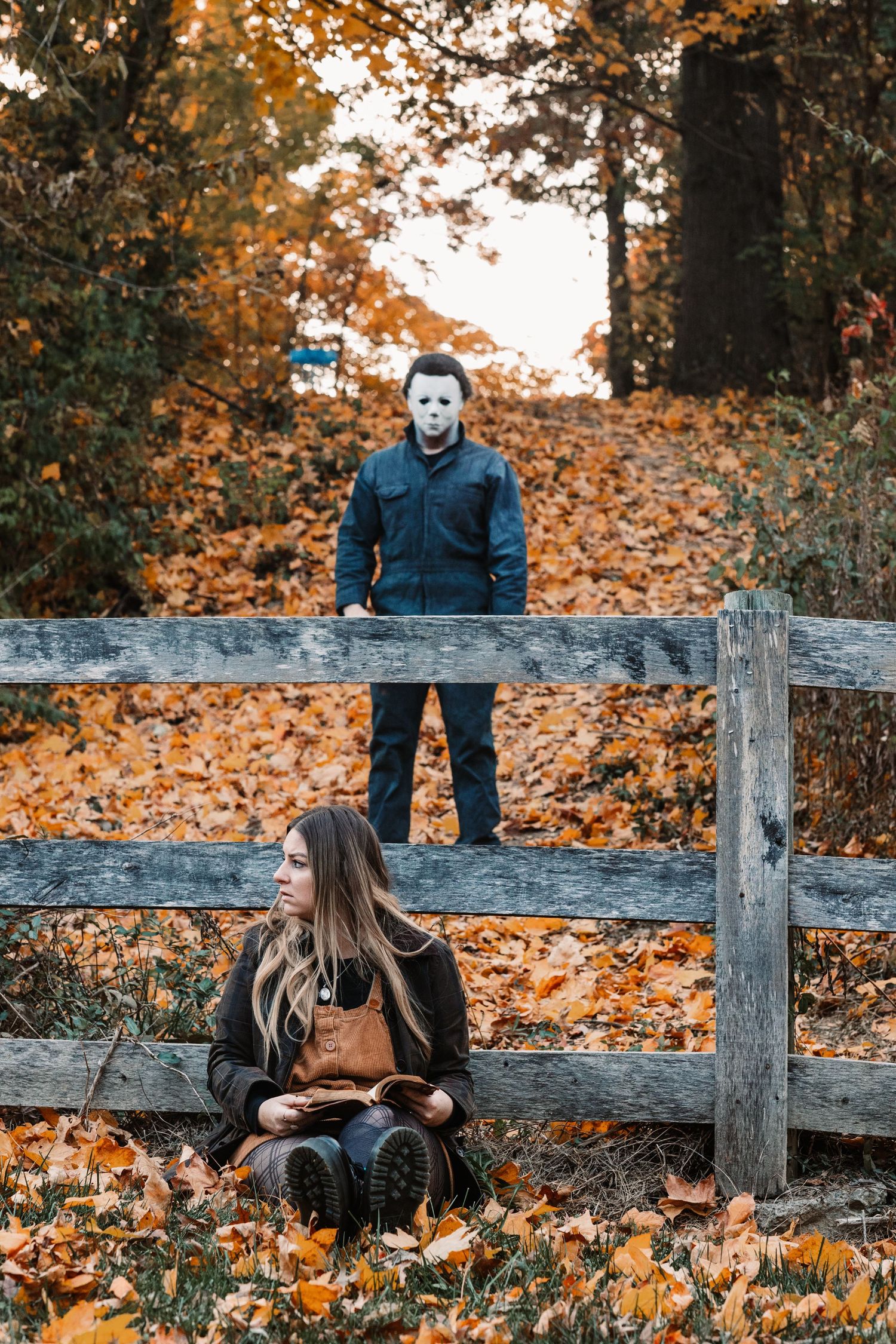 Woman sits by rustic fence unaware of man in Michael Myers Halloween costume standing behind her in autumn park.