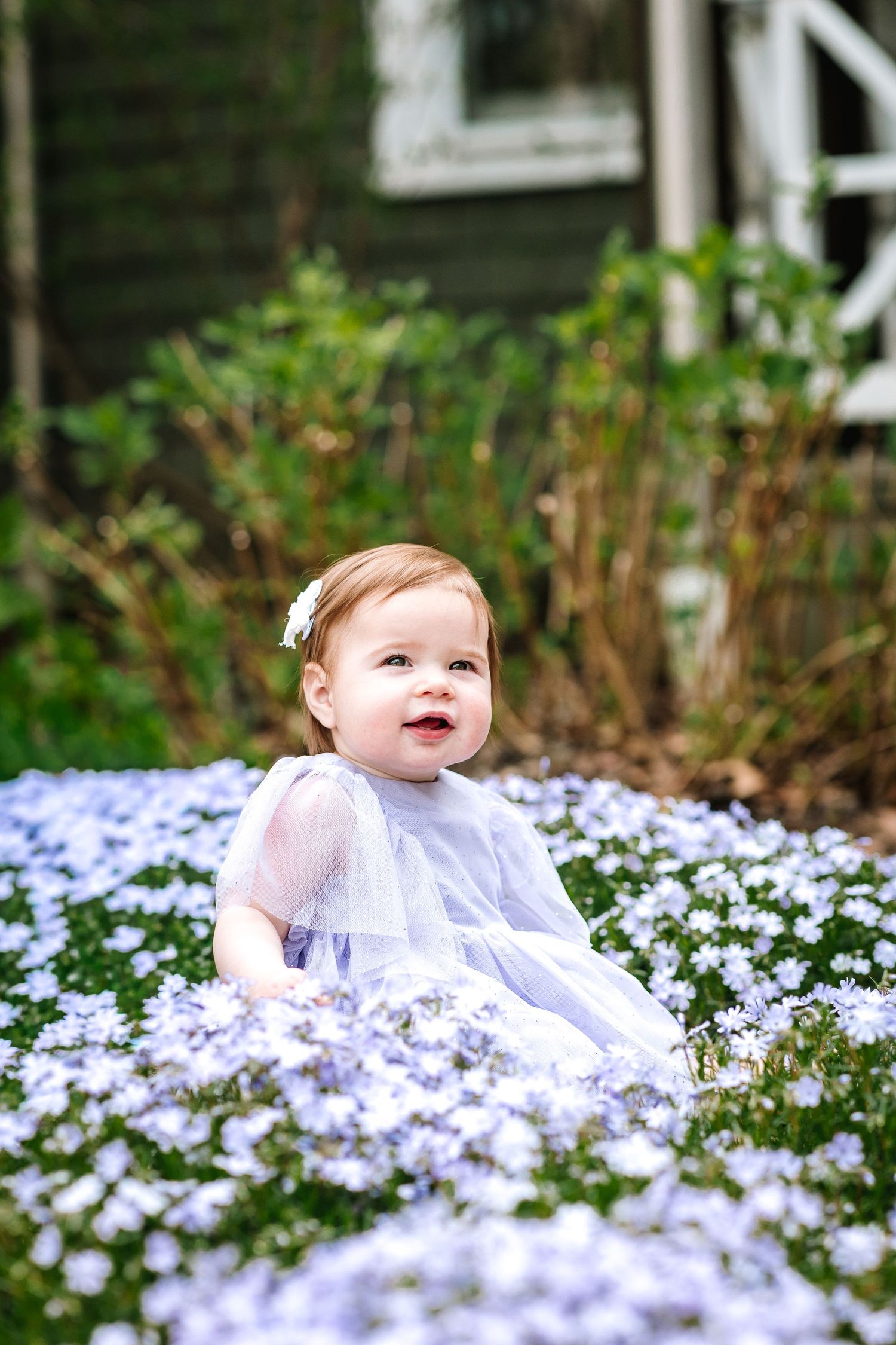 Adorable baby girl in white dress sitting among purple wildflowers in a garden.