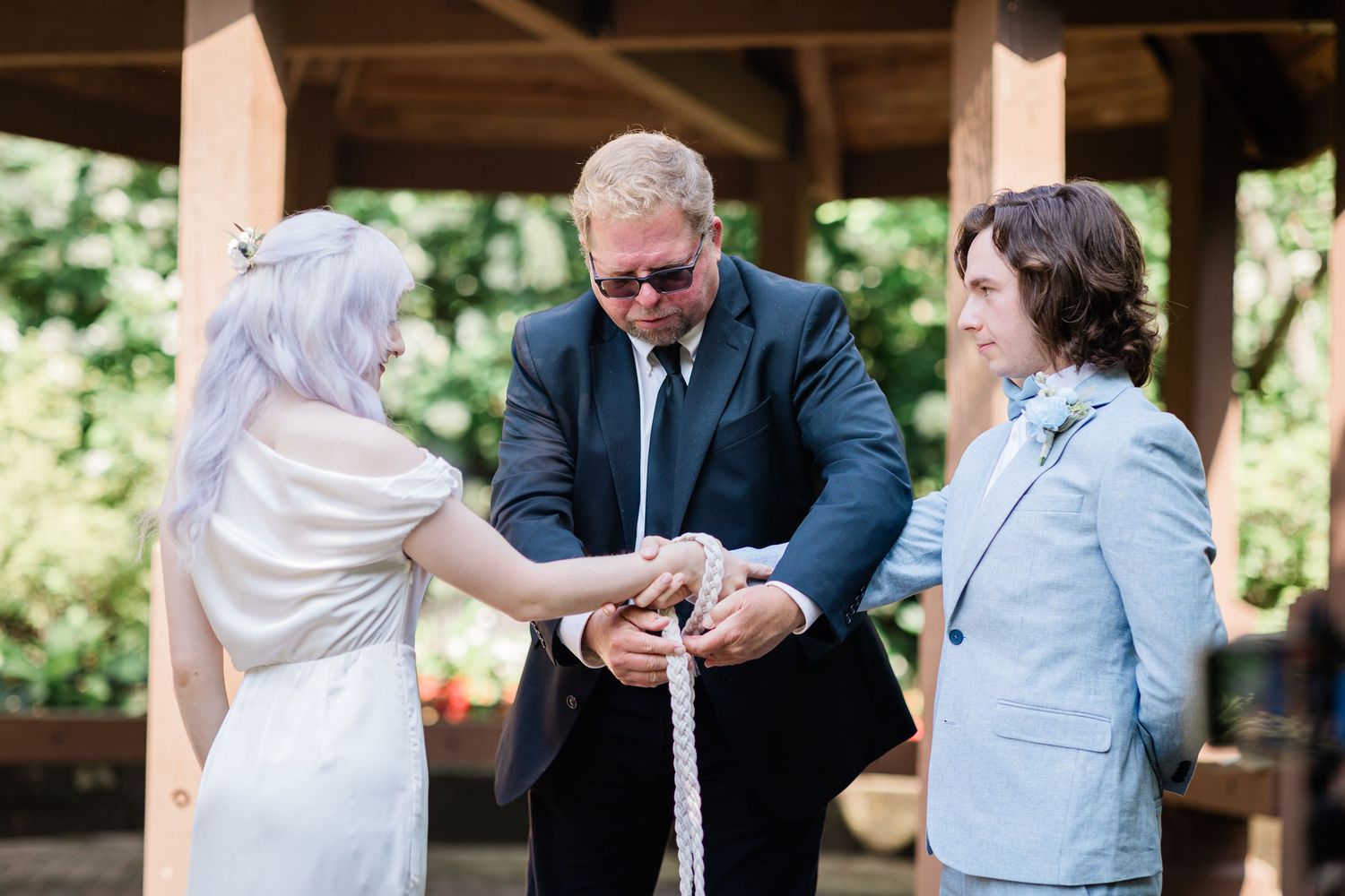 Officiant performs rope handfasting ceremony with bride in white and groom in light blue suit outdoors.
