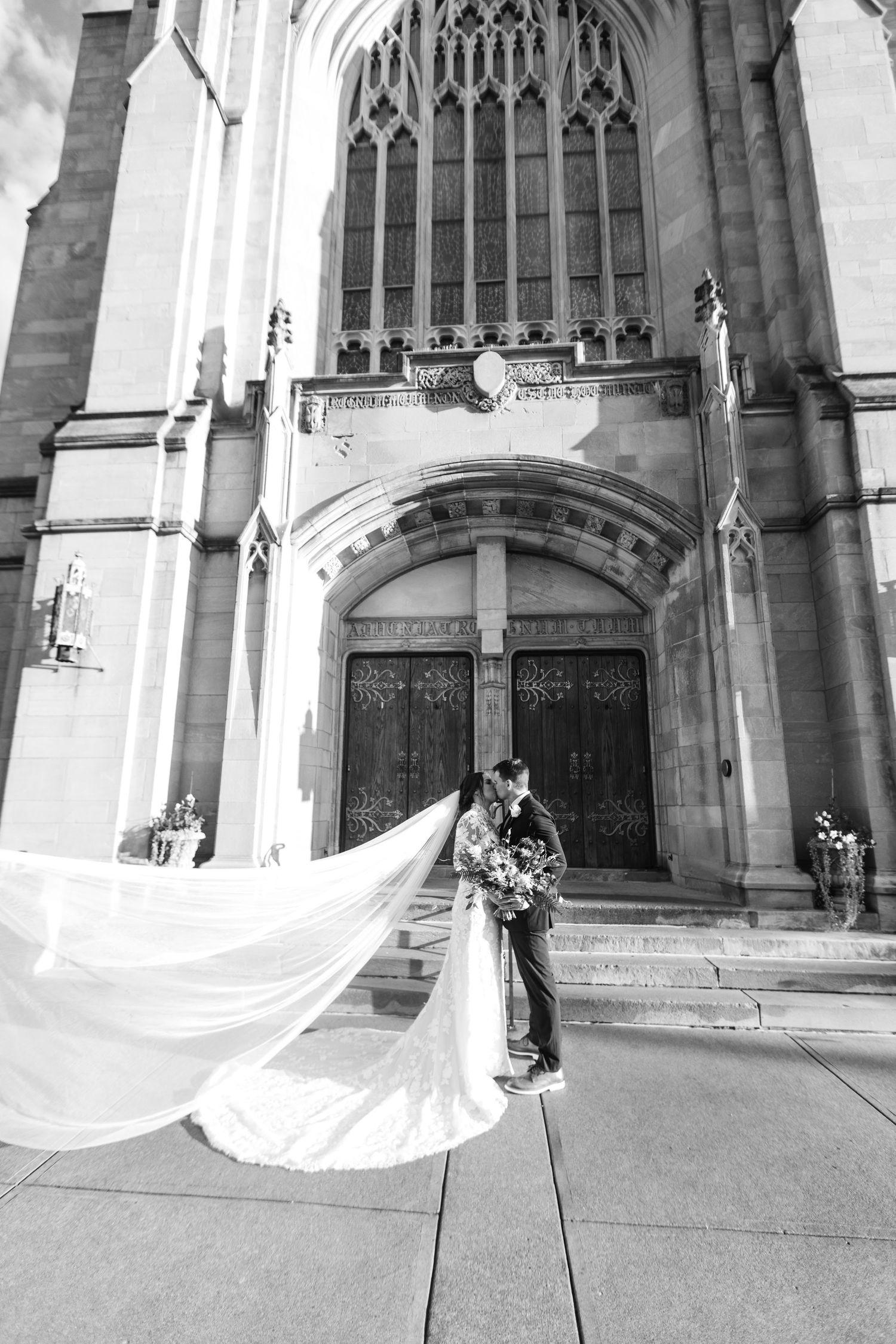 Bride and groom kiss outside grand Gothic church entrance as dramatic cathedral-length veil billows in wind.