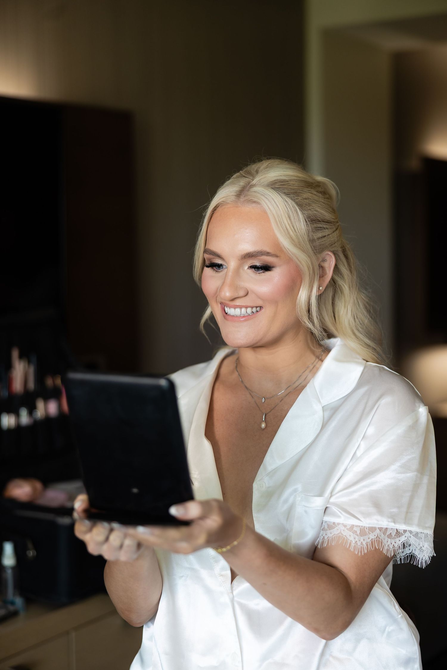 Smiling blonde bride in white robe checking her makeup in a mirror while getting ready.