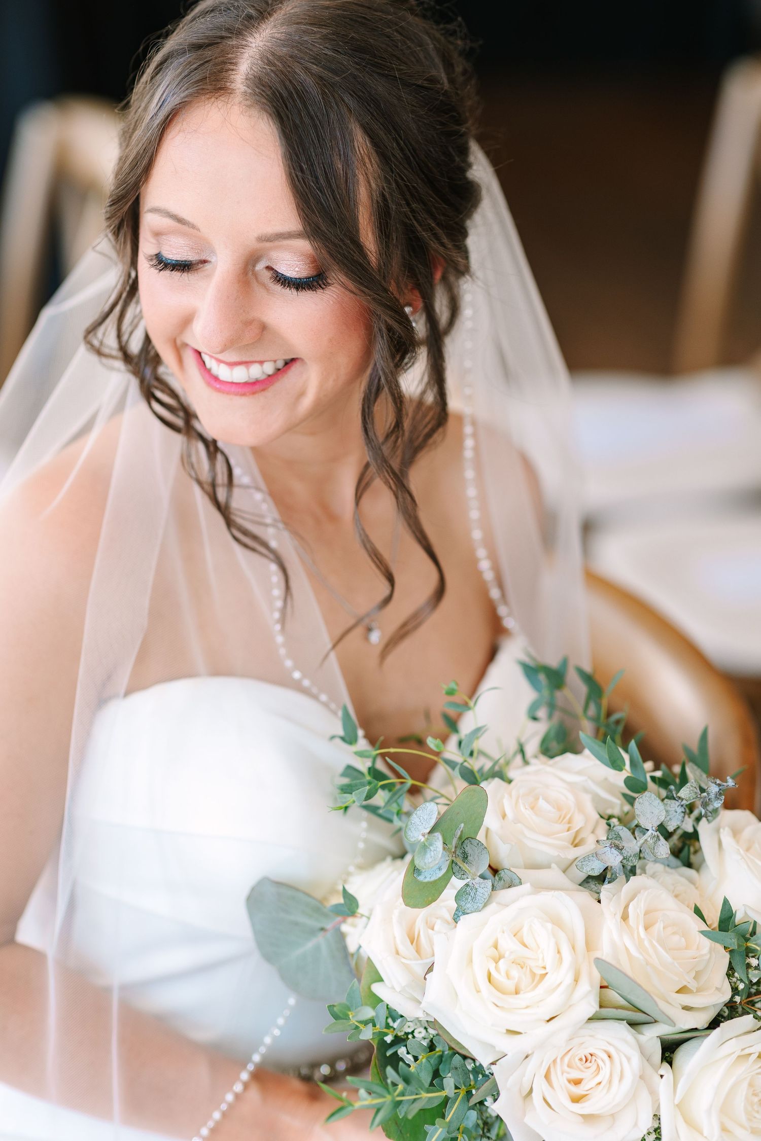 Smiling bride in a white strapless gown holding a bouquet of white roses with a pearl-edged veil.