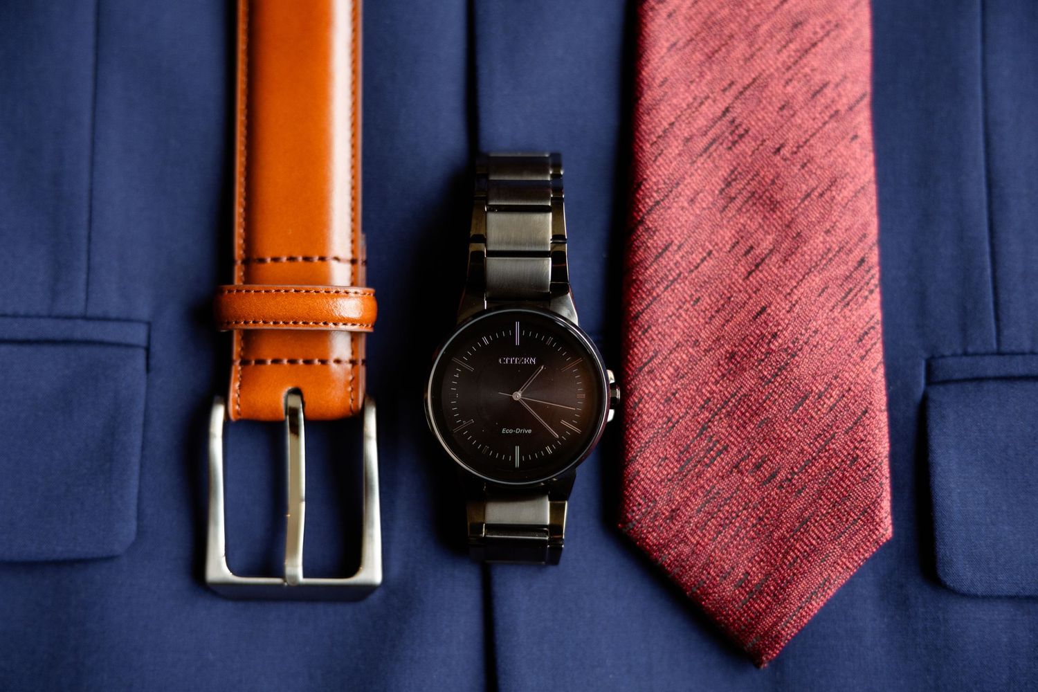 Groom's accessories flat lay featuring a brown leather belt, black watch, and red tie on a blue suit.