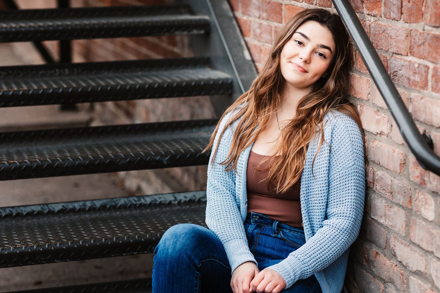 Young woman in blue cardigan sitting on steps beside a brick wall in a warm outdoor setting.