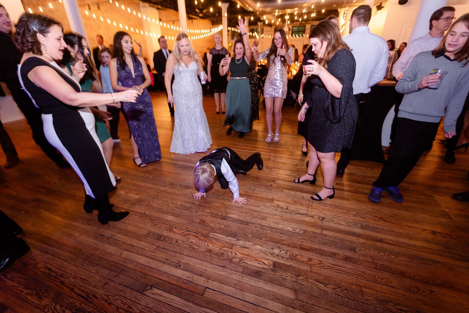 Wedding reception guests laughing as a toddler crawls on the dance floor beneath string lights in a rustic venue.