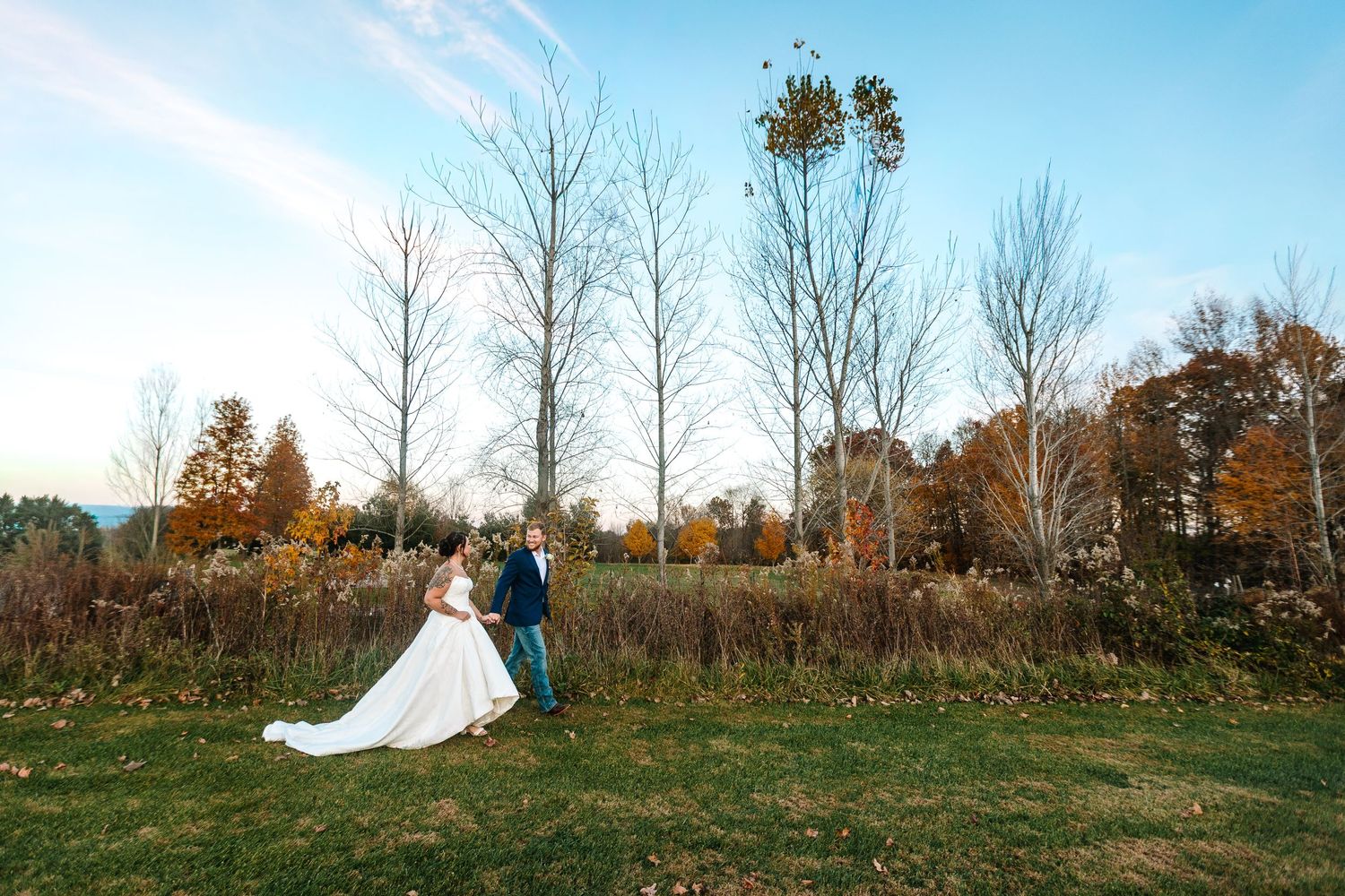 Bride and groom walk hand in hand through a scenic autumn landscape with colorful fall foliage.