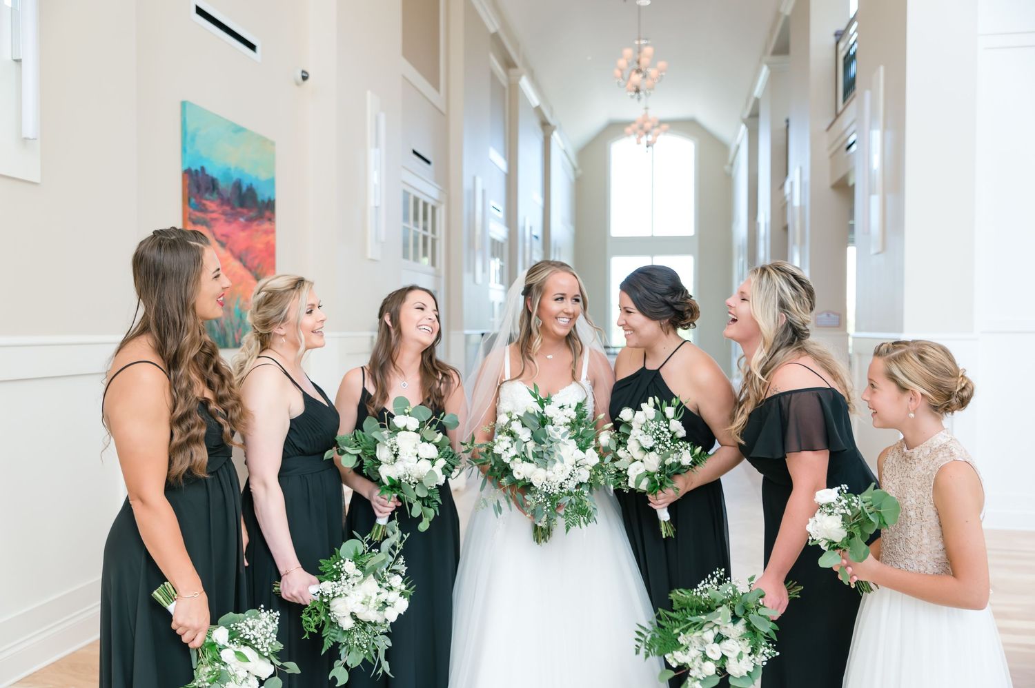 Bride with bridesmaids in black dresses holding white and greenery bouquets in elegant hallway at Cooper Creek.