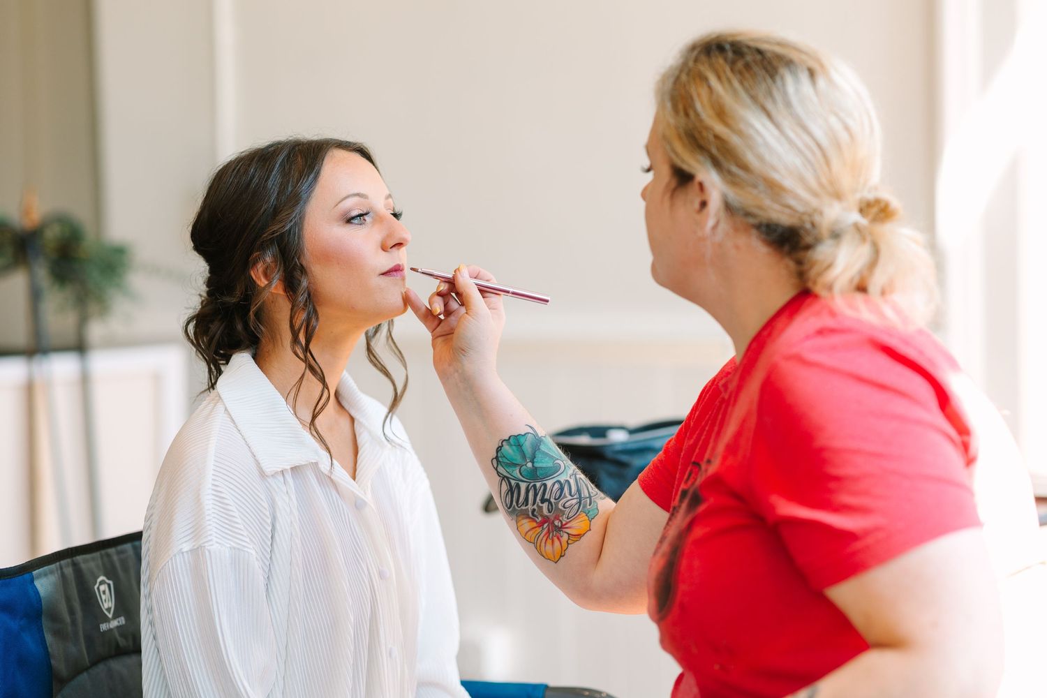 Makeup artist with tattoos applying lipstick to a bride in a white shirt before her wedding day.