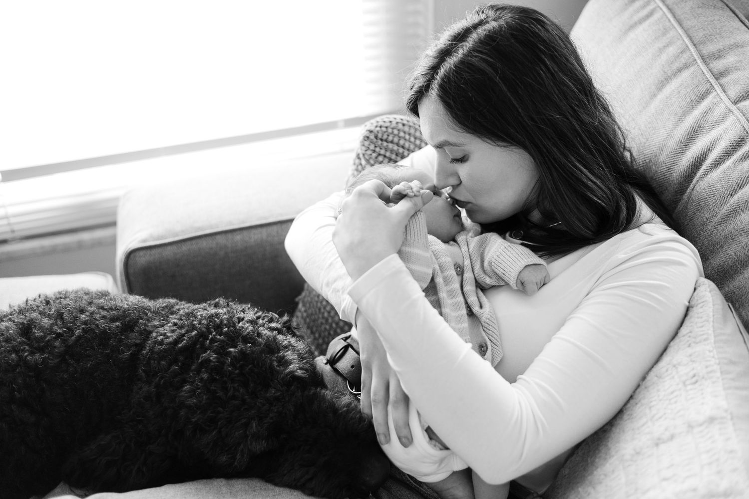 Black and white photo of a mother tenderly kissing her newborn baby on a couch with a dog nearby.