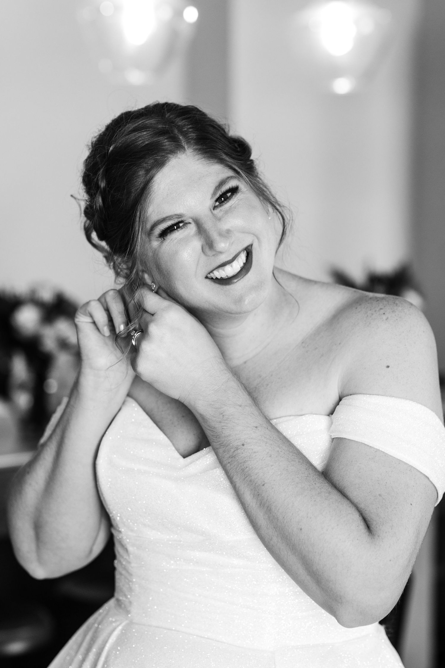 Smiling bride in an off-shoulder white gown adjusting her earrings in a black and white portrait photo.