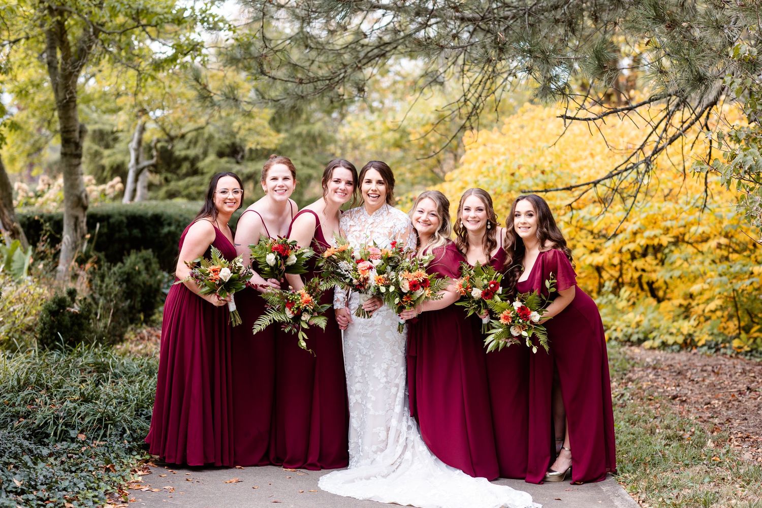 Bride and six bridesmaids in burgundy gowns pose with colorful bouquets in fall garden setting.