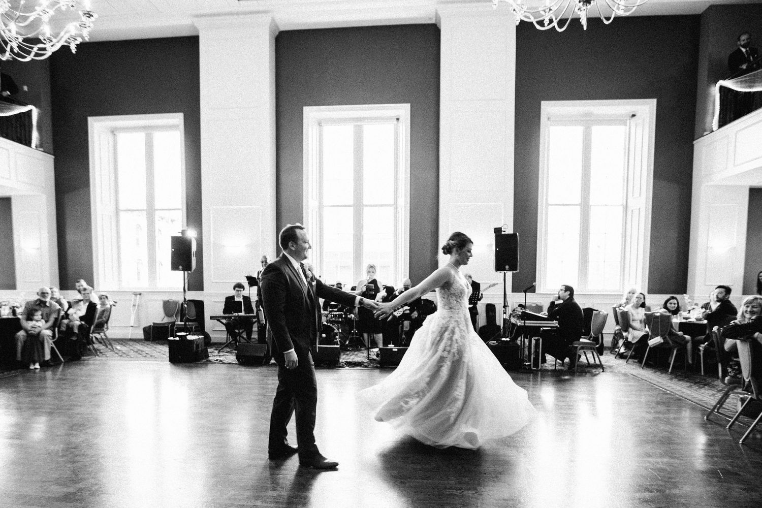 Bride and groom share their first dance in an elegant ballroom with tall windows and wedding guests seated nearby.