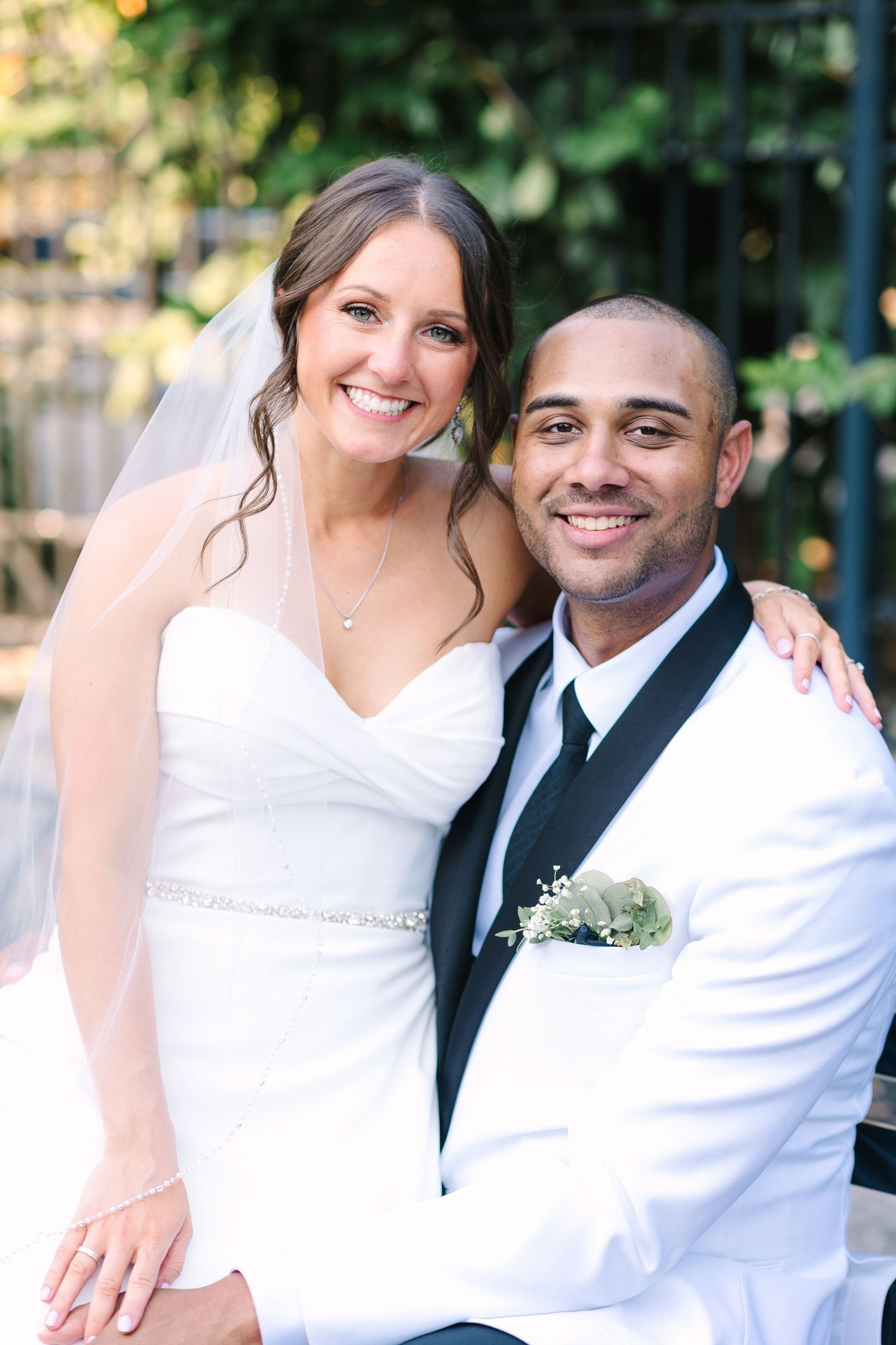 Smiling bride in white gown and groom in white tuxedo posing together outdoors in garden setting.