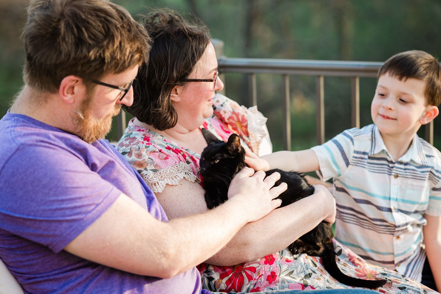 A family of three laughs together on a porch while holding a black cat.