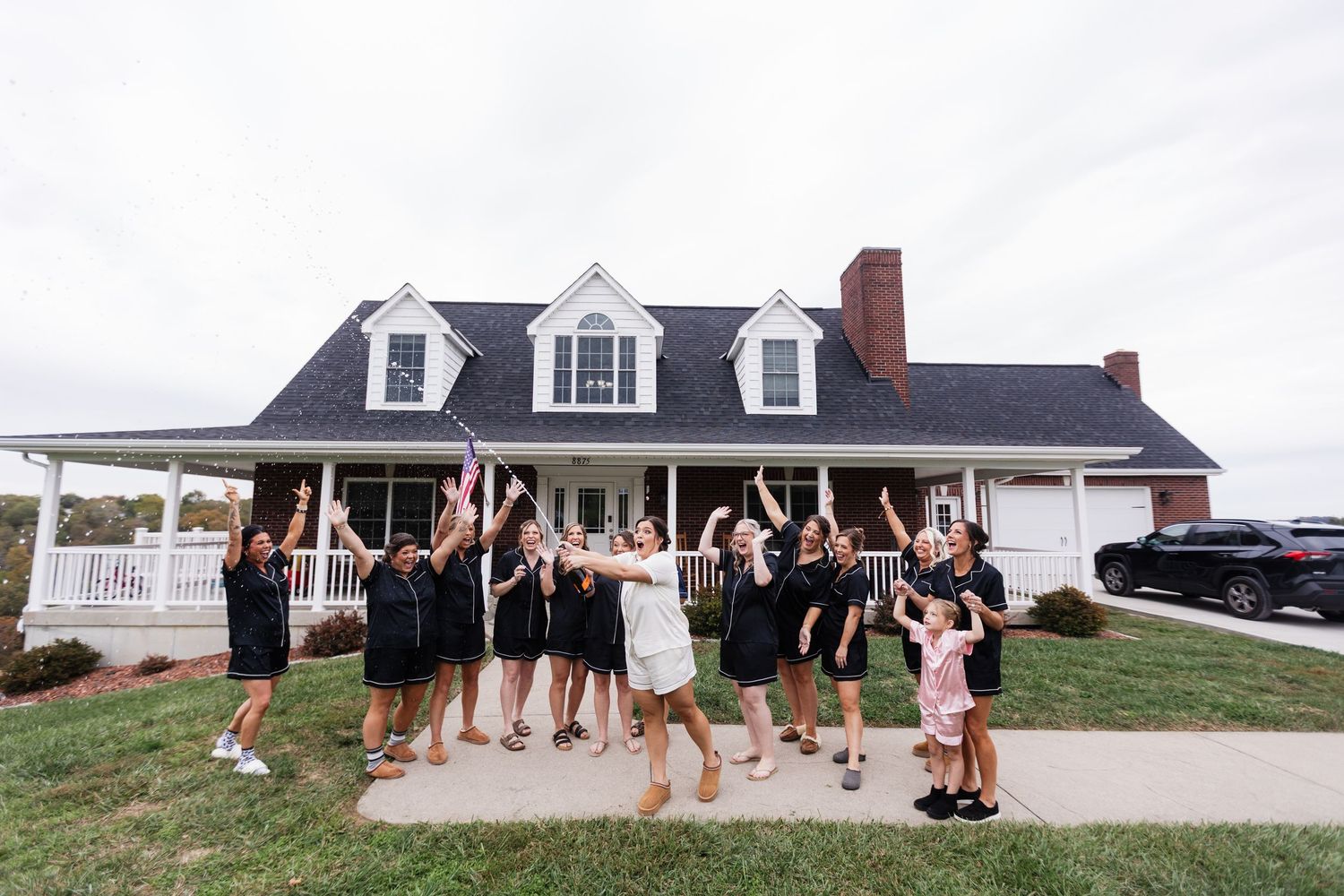 Bridal party in matching black outfits celebrating outside a white farmhouse.