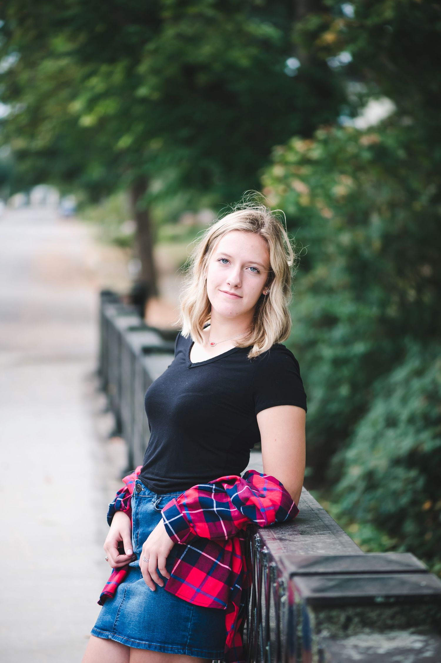 Blonde teenage girl leans on a wooden fence railing wearing a black tee and plaid shirt tied at waist.