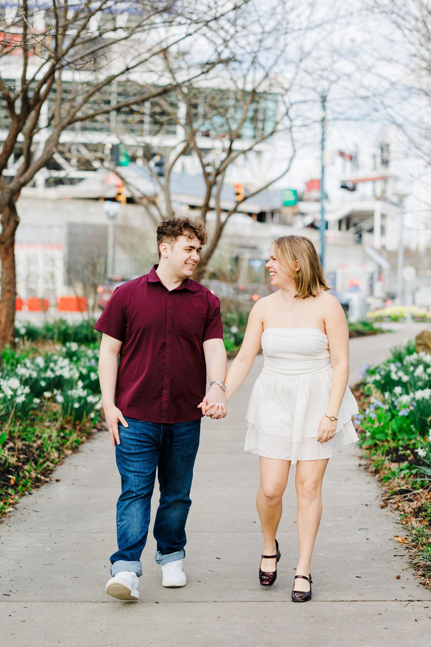 A smiling couple walks hand in hand along a city sidewalk lined with spring flowers and bare trees.