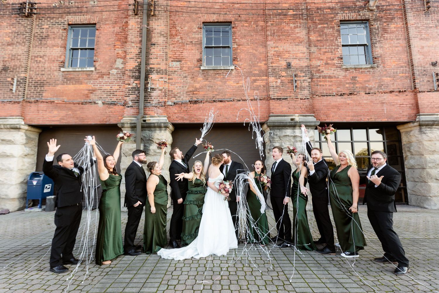 Large wedding party celebrating in front of a rustic brick building, bridesmaids in emerald green gowns.