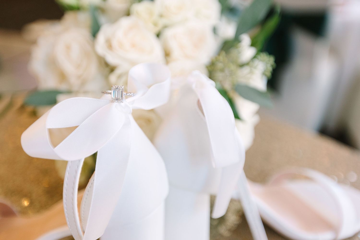 White wedding ring box adorned with satin ribbon bow beside white rose bridal bouquet on gold table.