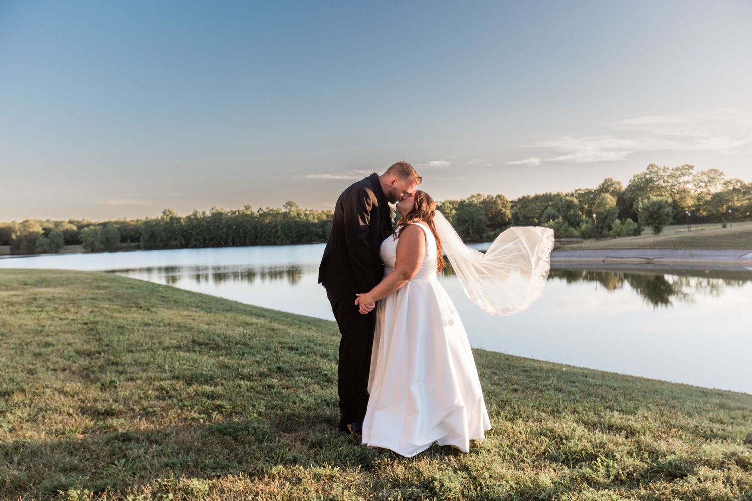 Bride and groom kiss romantically by a serene lake at golden hour with veil flowing at Lake Lyndsay.