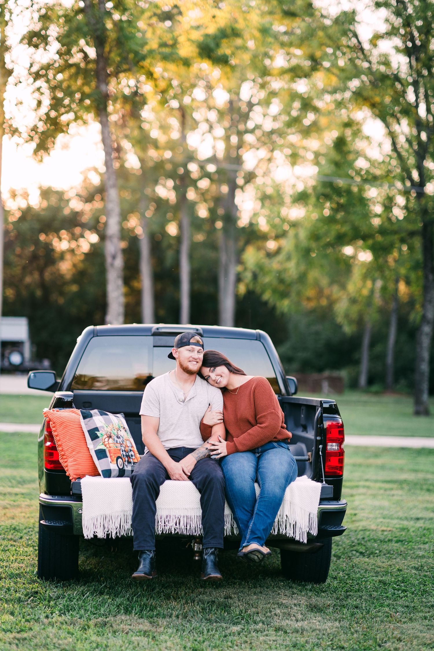 A couple sits closely together on a truck tailgate surrounded by cozy blankets and pillows at sunset.