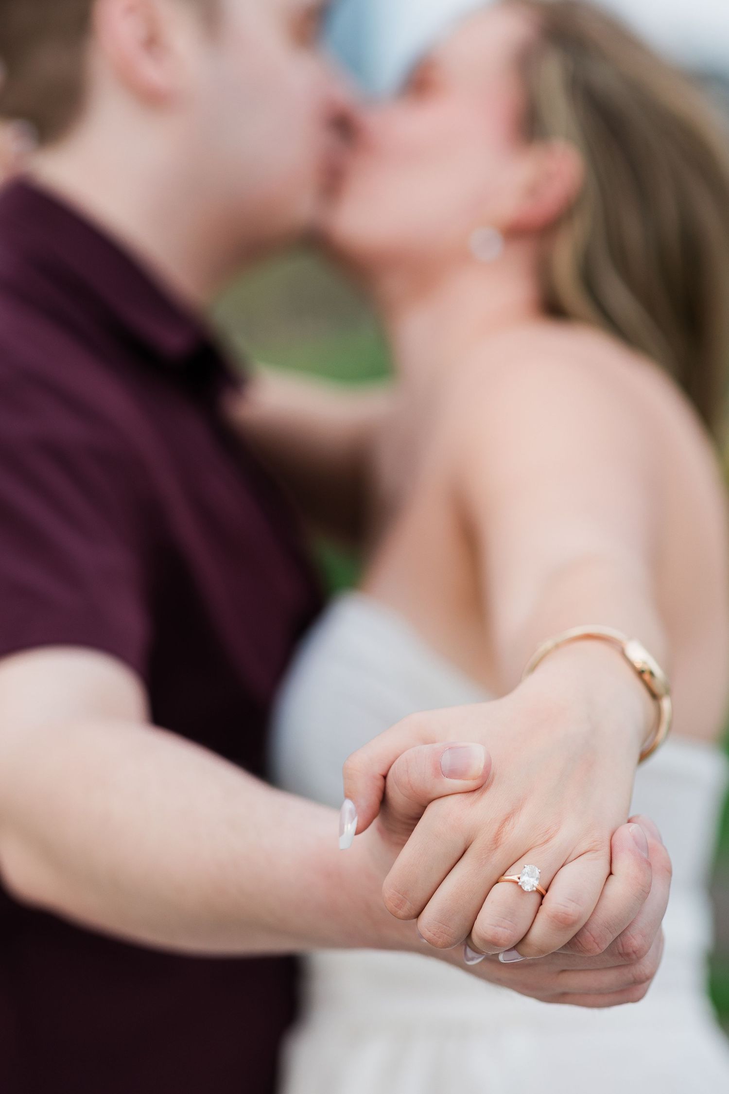 An engaged couple kisses while displaying a sparkling diamond engagement ring on the womans hand in the foreground.
