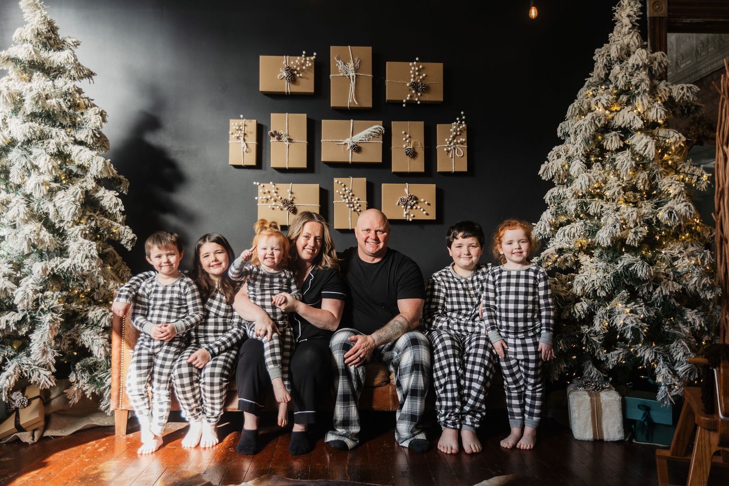 A large family in matching plaid pajamas poses between two flocked Christmas trees with wrapped gifts.