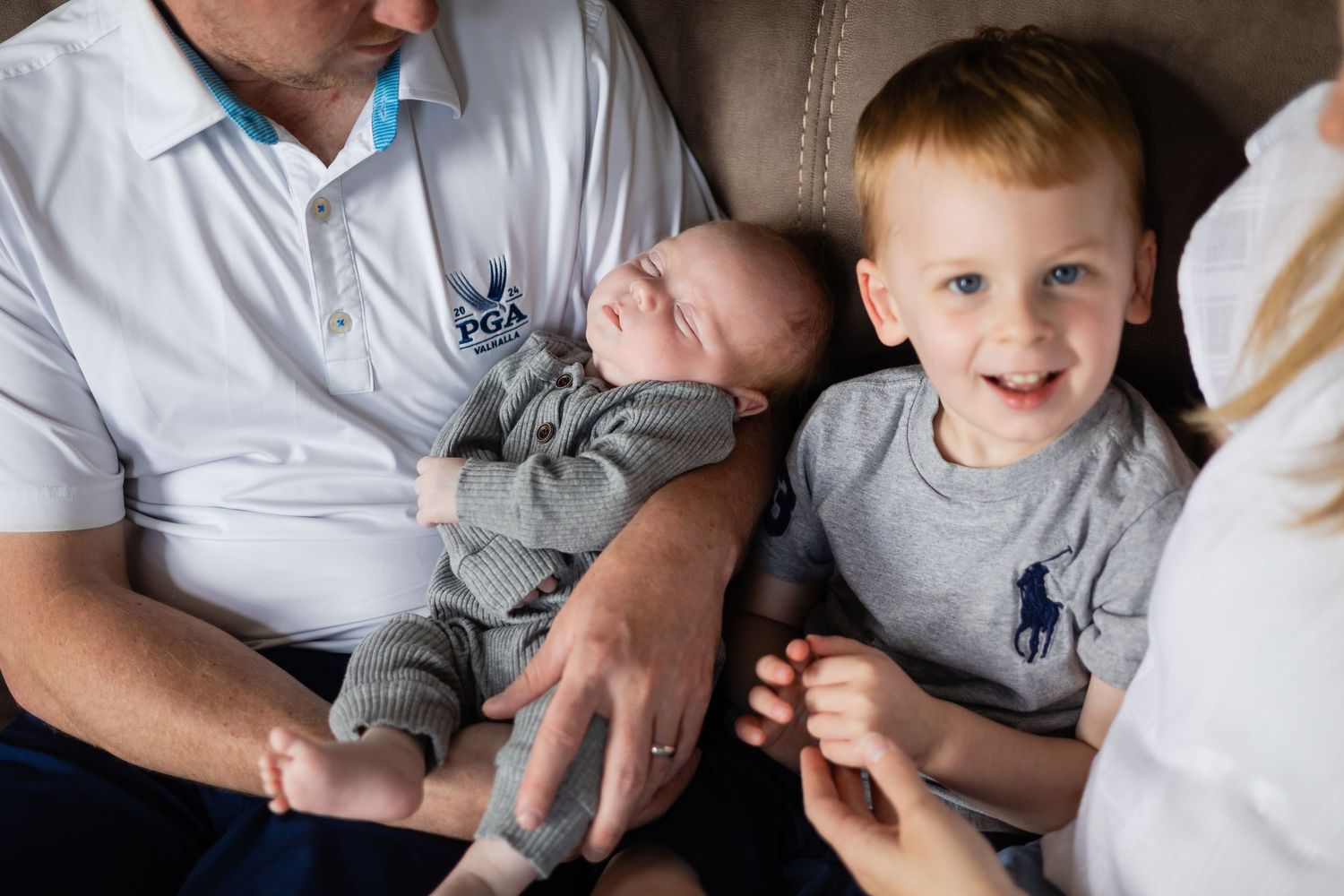 A young red-haired boy smiles proudly beside his sleeping newborn sibling held by their father.